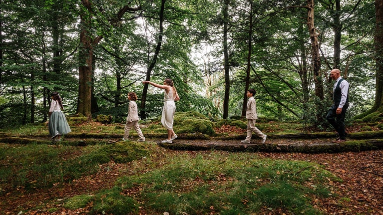 Bride and groom accompanied by three children are walking along a lush green woodland path.